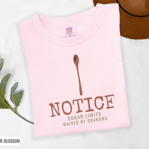 Blossom Comfort Colors t-shirt printed with the words Notice Sugar Limits Waived By Grandma and a drawing of a painted wooden spoon crossed over a cinnamon stick bundle, folded flat lay