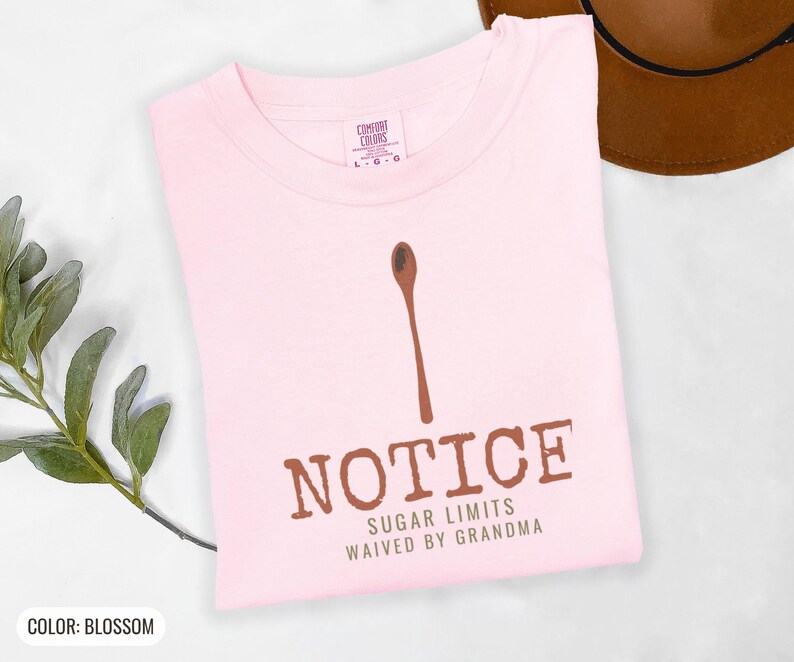 Blossom Comfort Colors t-shirt printed with the words Notice Sugar Limits Waived By Grandma and a drawing of a painted wooden spoon crossed over a cinnamon stick bundle, folded flat lay