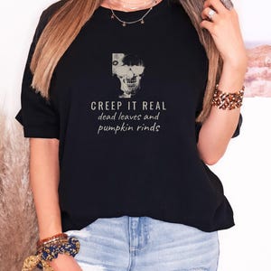 Black Comfort Colors t-shirt printed with the words Creep It Real Dead Leaves And Pumpkin Rinds and a drawing of a grinning skull with black sunglasses, worn by a woman
