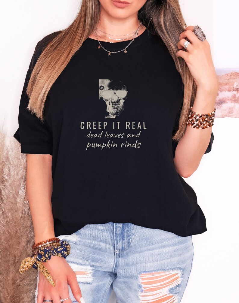 Black Comfort Colors t-shirt printed with the words Creep It Real Dead Leaves And Pumpkin Rinds and a drawing of a grinning skull with black sunglasses, worn by a woman