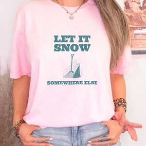 Blossom Comfort Colors t-shirt printed with the words Let It Snow Somewhere Else and a drawing of a metal snow shovel propped beside an empty bag of rock salt, worn by a woman