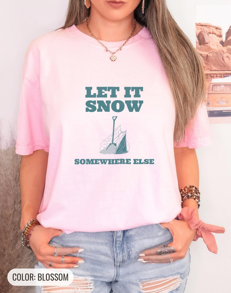 Blossom Comfort Colors t-shirt printed with the words Let It Snow Somewhere Else and a drawing of a metal snow shovel propped beside an empty bag of rock salt, worn by a woman