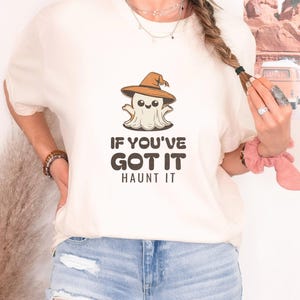 Ivory Comfort Colors t-shirt printed with the words If You'Ve Got It Haunt It and a drawing of a cute retro cartoon ghost wearing a witch hat, worn by a woman