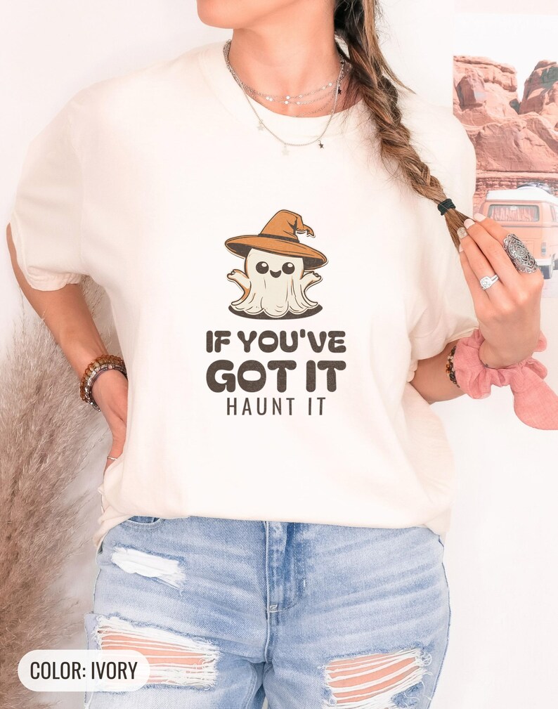 Ivory Comfort Colors t-shirt printed with the words If You'Ve Got It Haunt It and a drawing of a cute retro cartoon ghost wearing a witch hat, worn by a woman
