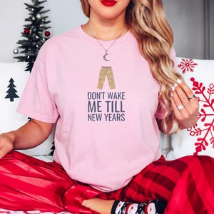 Blossom Comfort Colors t-shirt printed with the words Don'T Wake Me Till New Years and a drawing of a pair of plaid pajama pants folded neatly on a wooden stool, worn by a woman