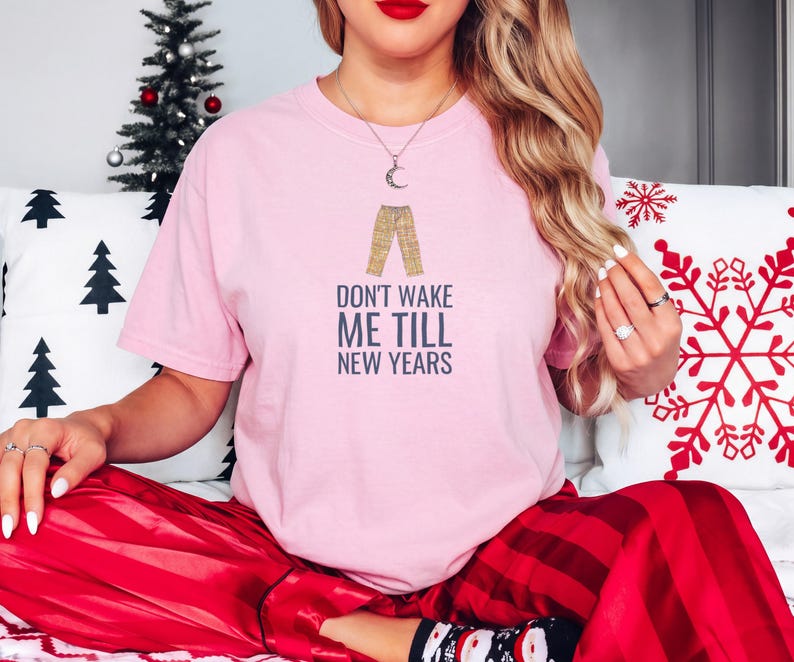 Blossom Comfort Colors t-shirt printed with the words Don'T Wake Me Till New Years and a drawing of a pair of plaid pajama pants folded neatly on a wooden stool, worn by a woman