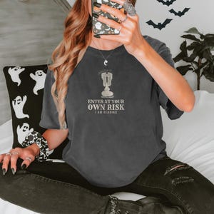 Pepper Comfort Colors t-shirt printed with the words Enter At Your Own Risk I Am Reading and a drawing of a brass candlestick with a single burning candle dripping wax, worn by a woman