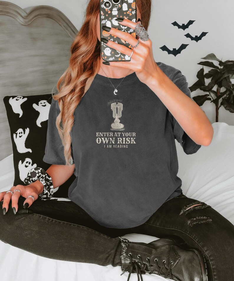 Pepper Comfort Colors t-shirt printed with the words Enter At Your Own Risk I Am Reading and a drawing of a brass candlestick with a single burning candle dripping wax, worn by a woman