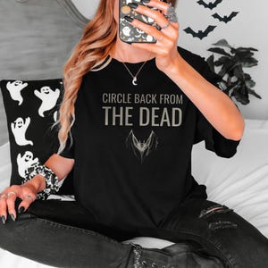 Black Comfort Colors t-shirt printed with the words Circle Back From The Dead and a drawing of a vintage silver paperclip bent into the shape of a bat, worn by a woman