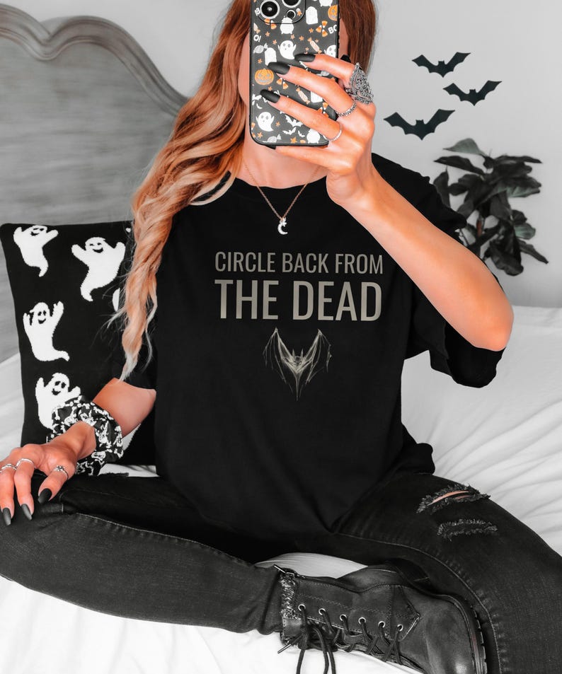 Black Comfort Colors t-shirt printed with the words Circle Back From The Dead and a drawing of a vintage silver paperclip bent into the shape of a bat, worn by a woman