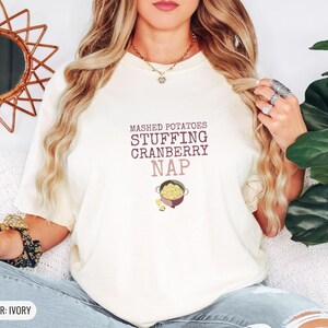 Ivory Comfort Colors t-shirt printed with the words Mashed Potatoes Stuffing Cranberry Nap and a drawing of a heavy enamel-coated cast iron Dutch oven filled with creamy mashed potatoes, steam lines, worn by a woman