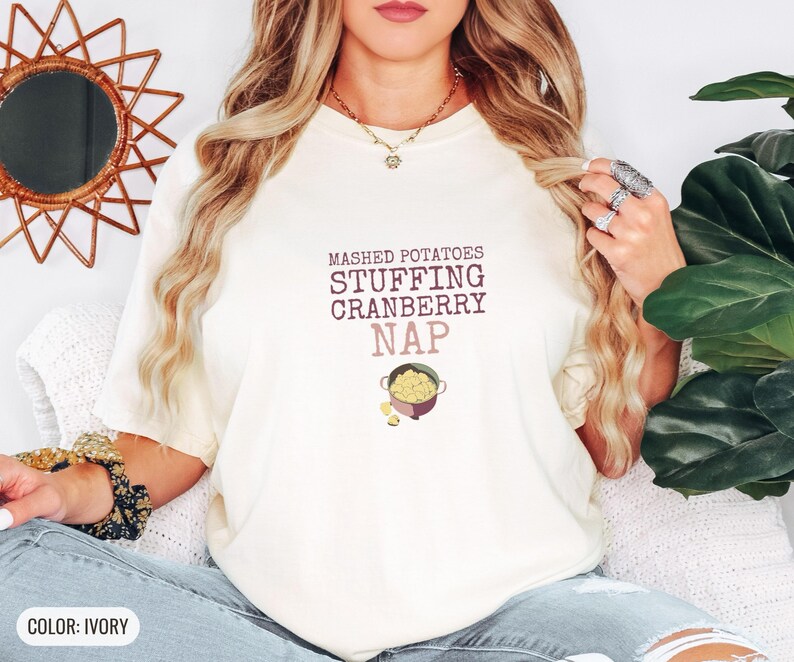 Ivory Comfort Colors t-shirt printed with the words Mashed Potatoes Stuffing Cranberry Nap and a drawing of a heavy enamel-coated cast iron Dutch oven filled with creamy mashed potatoes, steam lines, worn by a woman