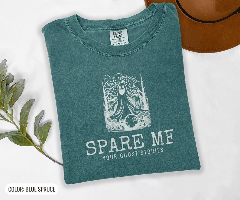 Blue Spruce Comfort Colors t-shirt printed with the words Spare Me Your Ghost Stories and a drawing of a friendly sheet ghost rolling a bowling ball towards a set of bowling pins, folded flat lay