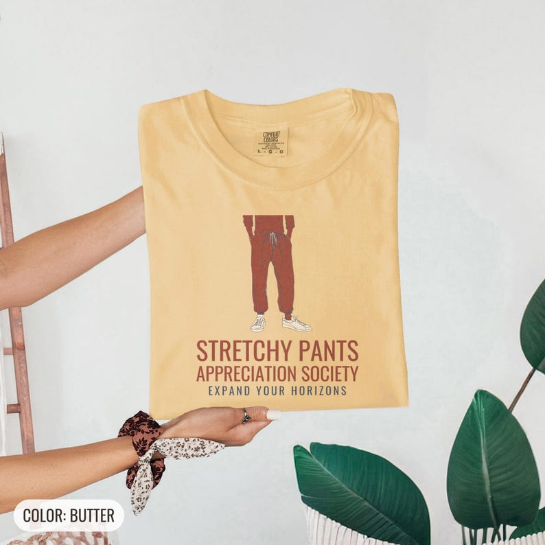 Butter Comfort Colors t-shirt printed with the words Stretchy Pants Appreciation Society Expand Your Horizons and a drawing of a pair of classic grey sweatpants with a visible drawstring, folded flat lay