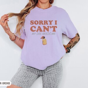 Orchid Comfort Colors t-shirt printed with the words Sorry I Can'T My Dog Needs Me and a drawing of a sleeping dachshund wearing a patterned knit sweater, worn by a woman