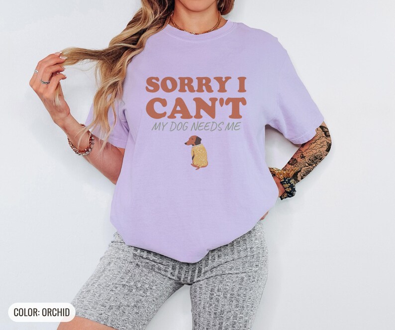 Orchid Comfort Colors t-shirt printed with the words Sorry I Can'T My Dog Needs Me and a drawing of a sleeping dachshund wearing a patterned knit sweater, worn by a woman