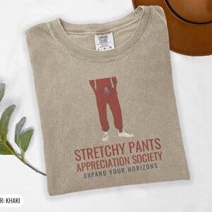 Khaki Comfort Colors t-shirt printed with the words Stretchy Pants Appreciation Society Expand Your Horizons and a drawing of a pair of classic grey sweatpants with a visible drawstring, folded flat lay