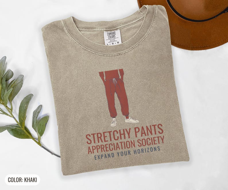 Khaki Comfort Colors t-shirt printed with the words Stretchy Pants Appreciation Society Expand Your Horizons and a drawing of a pair of classic grey sweatpants with a visible drawstring, folded flat lay