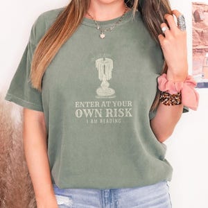 Moss Comfort Colors t-shirt printed with the words Enter At Your Own Risk I Am Reading and a drawing of a brass candlestick with a single burning candle dripping wax, worn by a woman