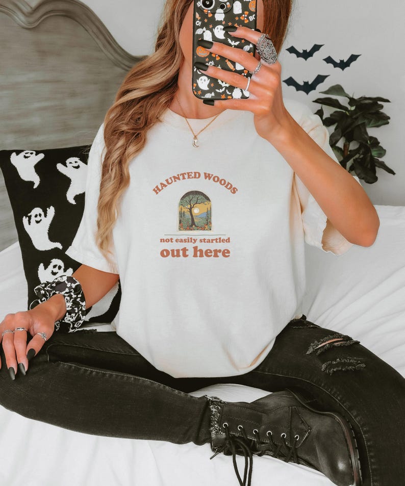 Ivory Comfort Colors t-shirt printed with the words Haunted Woods Not Easily Startled Out Here and a drawing of a dark forest of gnarled, leafless trees under a full moon, worn by a woman