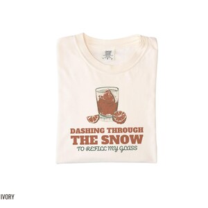 Ivory Comfort Colors t-shirt printed with the words Dashing Through The Snow To Refill My Glass and a drawing of a heavy crystal whiskey tumbler with two floating ice cubes beside a sprig of pine needles, folded flat lay