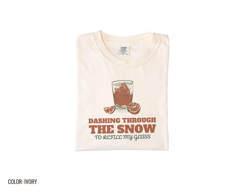 Ivory Comfort Colors t-shirt printed with the words Dashing Through The Snow To Refill My Glass and a drawing of a heavy crystal whiskey tumbler with two floating ice cubes beside a sprig of pine needles, folded flat lay