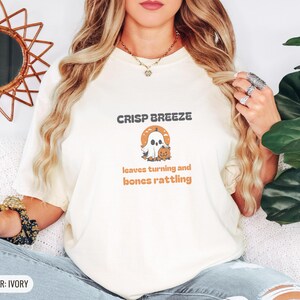 Ivory Comfort Colors t-shirt printed with the words Crisp Breeze Leaves Turning And Bones Rattling and a drawing of a large cute smiling ghost beside a carved jack-o-lantern, worn by a woman