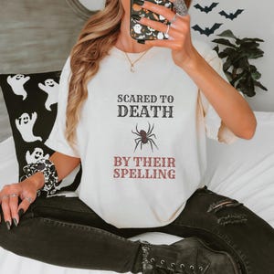 Ivory Comfort Colors t-shirt printed with the words Scared To Death By Their Spelling and a drawing of a highly detailed, crisp linocut spider sitting on top of a red-graded spelling test paper, worn by a woman