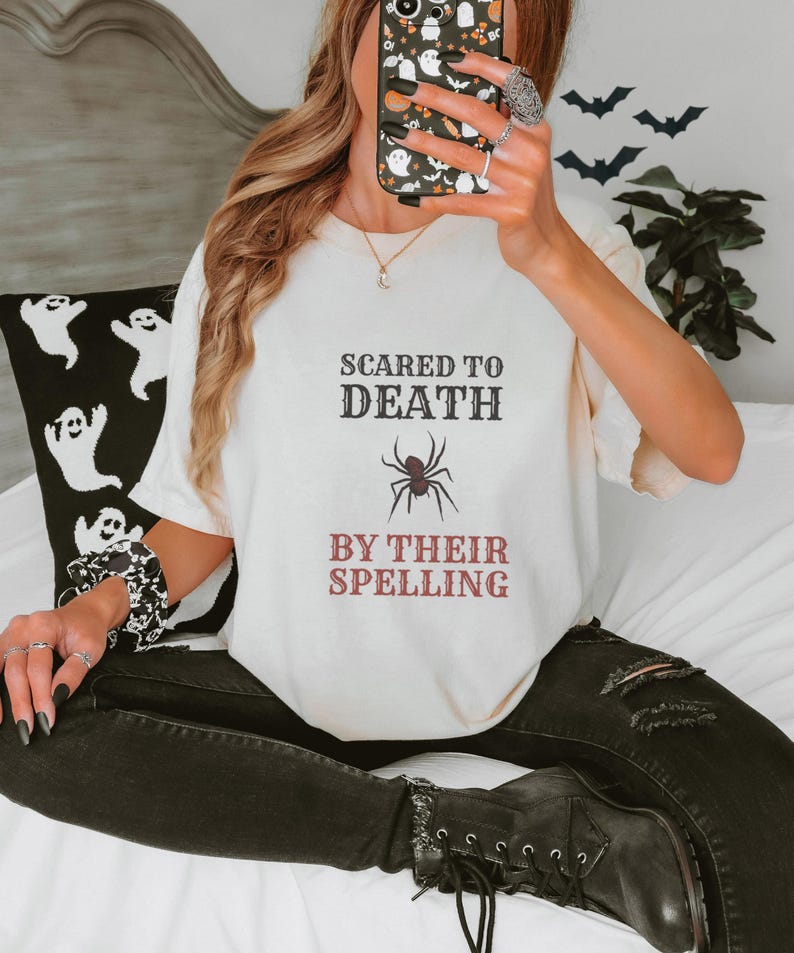 Ivory Comfort Colors t-shirt printed with the words Scared To Death By Their Spelling and a drawing of a highly detailed, crisp linocut spider sitting on top of a red-graded spelling test paper, worn by a woman