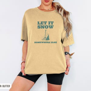 Butter Comfort Colors t-shirt printed with the words Let It Snow Somewhere Else and a drawing of a metal snow shovel propped beside an empty bag of rock salt, worn by a woman