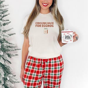 Ivory Comfort Colors t-shirt printed with the words Circling Back For Eggnog and a drawing of a vintage paper milk carton labeled eggnog with a green pine needle design on the side, worn by a woman