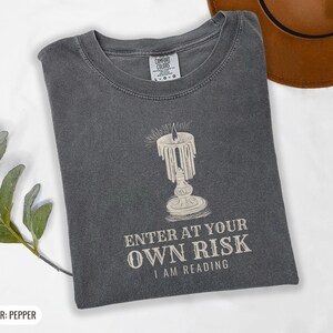 Pepper Comfort Colors t-shirt printed with the words Enter At Your Own Risk I Am Reading and a drawing of a brass candlestick with a single burning candle dripping wax, folded flat lay