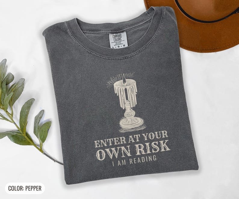 Pepper Comfort Colors t-shirt printed with the words Enter At Your Own Risk I Am Reading and a drawing of a brass candlestick with a single burning candle dripping wax, folded flat lay