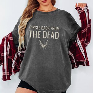 Pepper Comfort Colors t-shirt printed with the words Circle Back From The Dead and a drawing of a vintage silver paperclip bent into the shape of a bat, worn by a woman