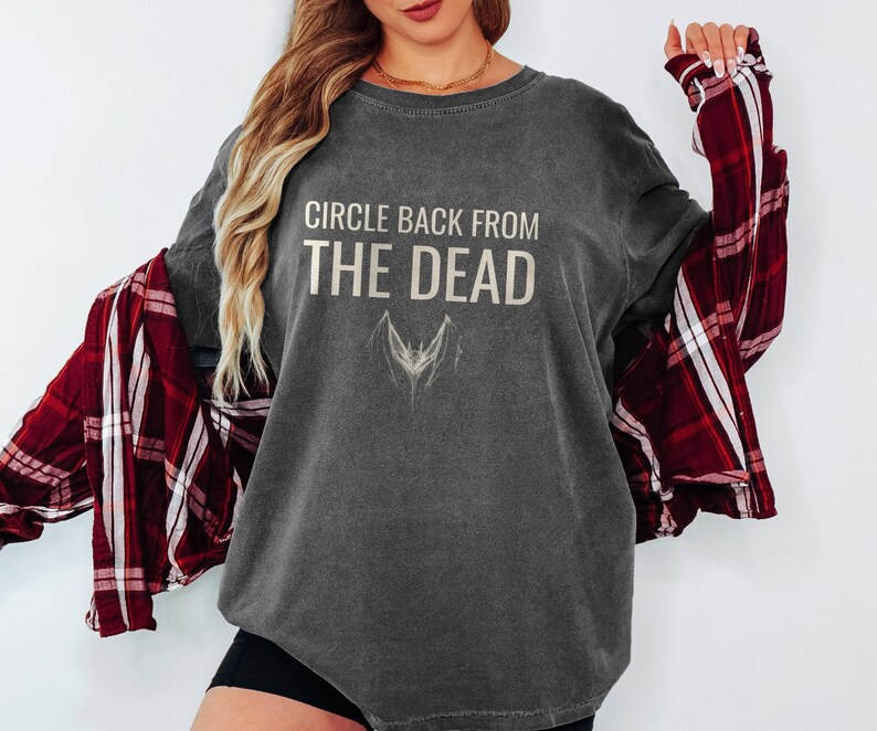 Pepper Comfort Colors t-shirt printed with the words Circle Back From The Dead and a drawing of a vintage silver paperclip bent into the shape of a bat, worn by a woman