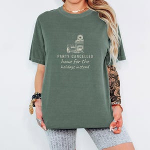 Moss Comfort Colors t-shirt printed with the words Party Cancelled Home For The Holidays Instead and a drawing of a cozy living room with a fireplace and Christmas decorations, a warm glow fills the space, worn by a woman