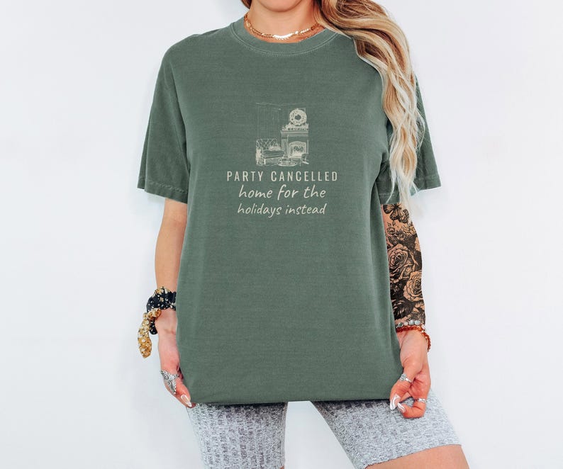 Moss Comfort Colors t-shirt printed with the words Party Cancelled Home For The Holidays Instead and a drawing of a cozy living room with a fireplace and Christmas decorations, a warm glow fills the space, worn by a woman