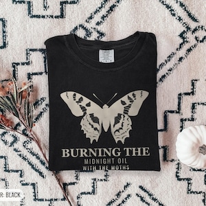 Black Comfort Colors t-shirt printed with the words Burning The Midnight Oil With The Moths and a drawing of a cecropia moth with feathery antennae, worn by a woman
