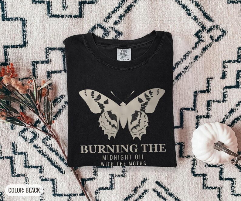 Black Comfort Colors t-shirt printed with the words Burning The Midnight Oil With The Moths and a drawing of a cecropia moth with feathery antennae, worn by a woman