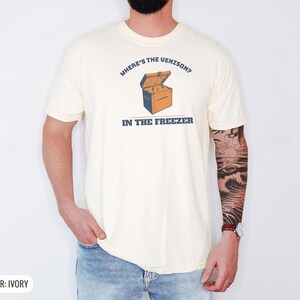 Ivory Comfort Colors t-shirt printed with the words Where'S The Venison? In The Freezer and a drawing of a vintage metal chest freezer with a pair of paper-wrapped butcher packages resting on top, worn by a man