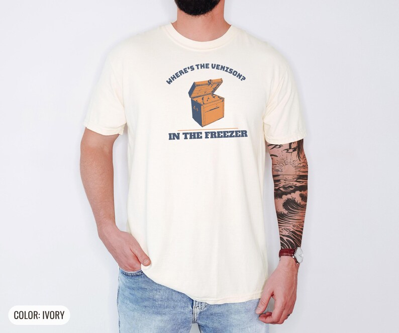 Ivory Comfort Colors t-shirt printed with the words Where'S The Venison? In The Freezer and a drawing of a vintage metal chest freezer with a pair of paper-wrapped butcher packages resting on top, worn by a man