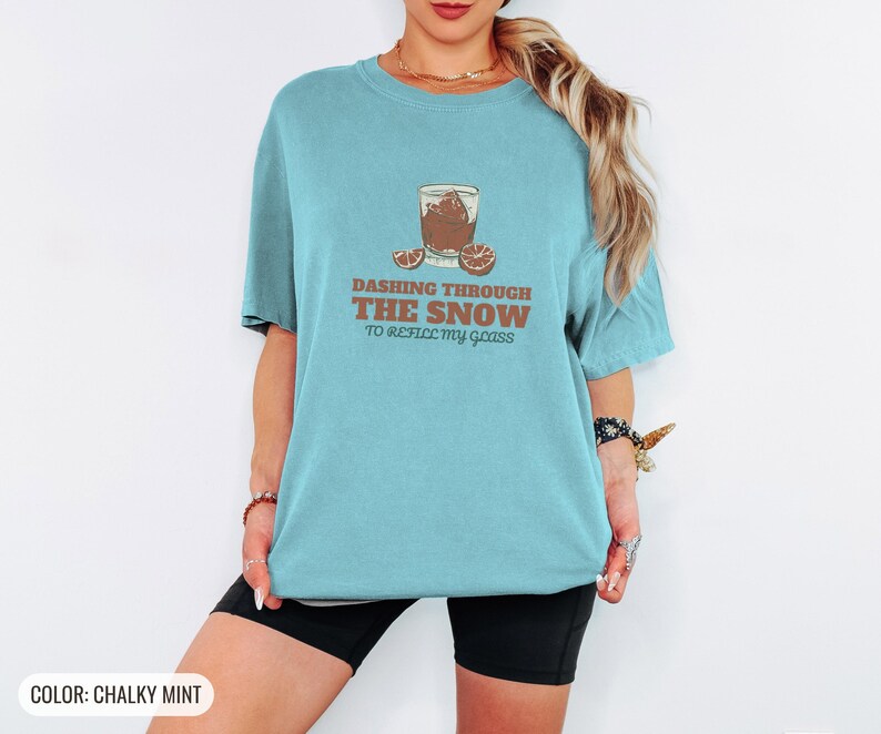 Chalky Mint Comfort Colors t-shirt printed with the words Dashing Through The Snow To Refill My Glass and a drawing of a heavy crystal whiskey tumbler with two floating ice cubes beside a sprig of pine needles, worn by a woman