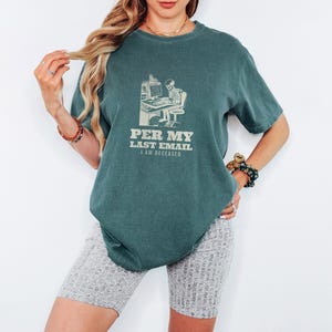 Blue Spruce Comfort Colors t-shirt printed with the words Per My Last Email I Am Deceased and a drawing of a skeleton sitting at an office desk typing on a computer keyboard, worn by a woman