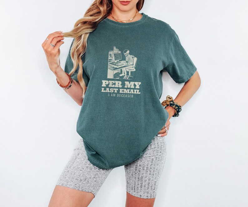 Blue Spruce Comfort Colors t-shirt printed with the words Per My Last Email I Am Deceased and a drawing of a skeleton sitting at an office desk typing on a computer keyboard, worn by a woman