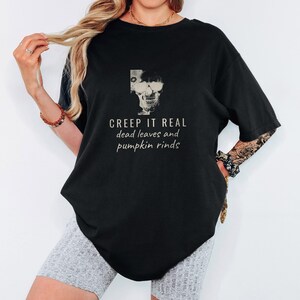 Black Comfort Colors t-shirt printed with the words Creep It Real Dead Leaves And Pumpkin Rinds and a drawing of a grinning skull with black sunglasses, worn by a woman