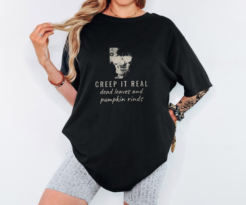 Black Comfort Colors t-shirt printed with the words Creep It Real Dead Leaves And Pumpkin Rinds and a drawing of a grinning skull with black sunglasses, worn by a woman