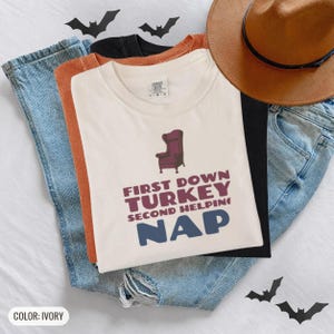 Ivory Comfort Colors t-shirt printed with the words First Down Turkey Second Helping Nap and a drawing of a vintage brown recliner with a footrest extended outward, worn by a woman