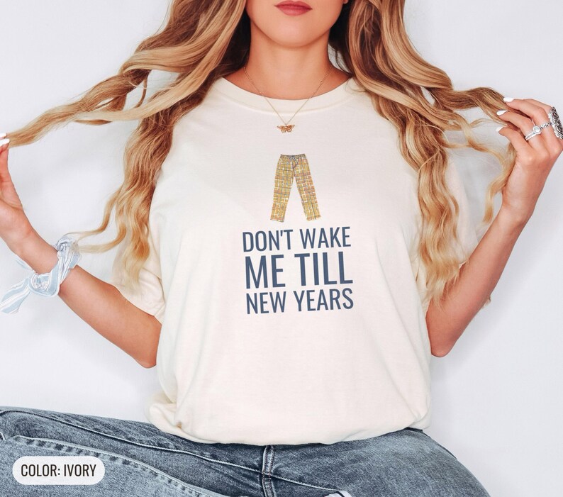 Ivory Comfort Colors t-shirt printed with the words Don'T Wake Me Till New Years and a drawing of a pair of plaid pajama pants folded neatly on a wooden stool, worn by a woman