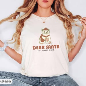 Ivory Comfort Colors t-shirt printed with the words Dear Santa The Ferret Did It and a drawing of a mischievous ferret dragging a striped Christmas stocking filled with shiny ornaments, worn by a woman