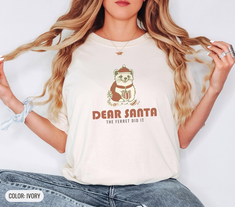 Ivory Comfort Colors t-shirt printed with the words Dear Santa The Ferret Did It and a drawing of a mischievous ferret dragging a striped Christmas stocking filled with shiny ornaments, worn by a woman